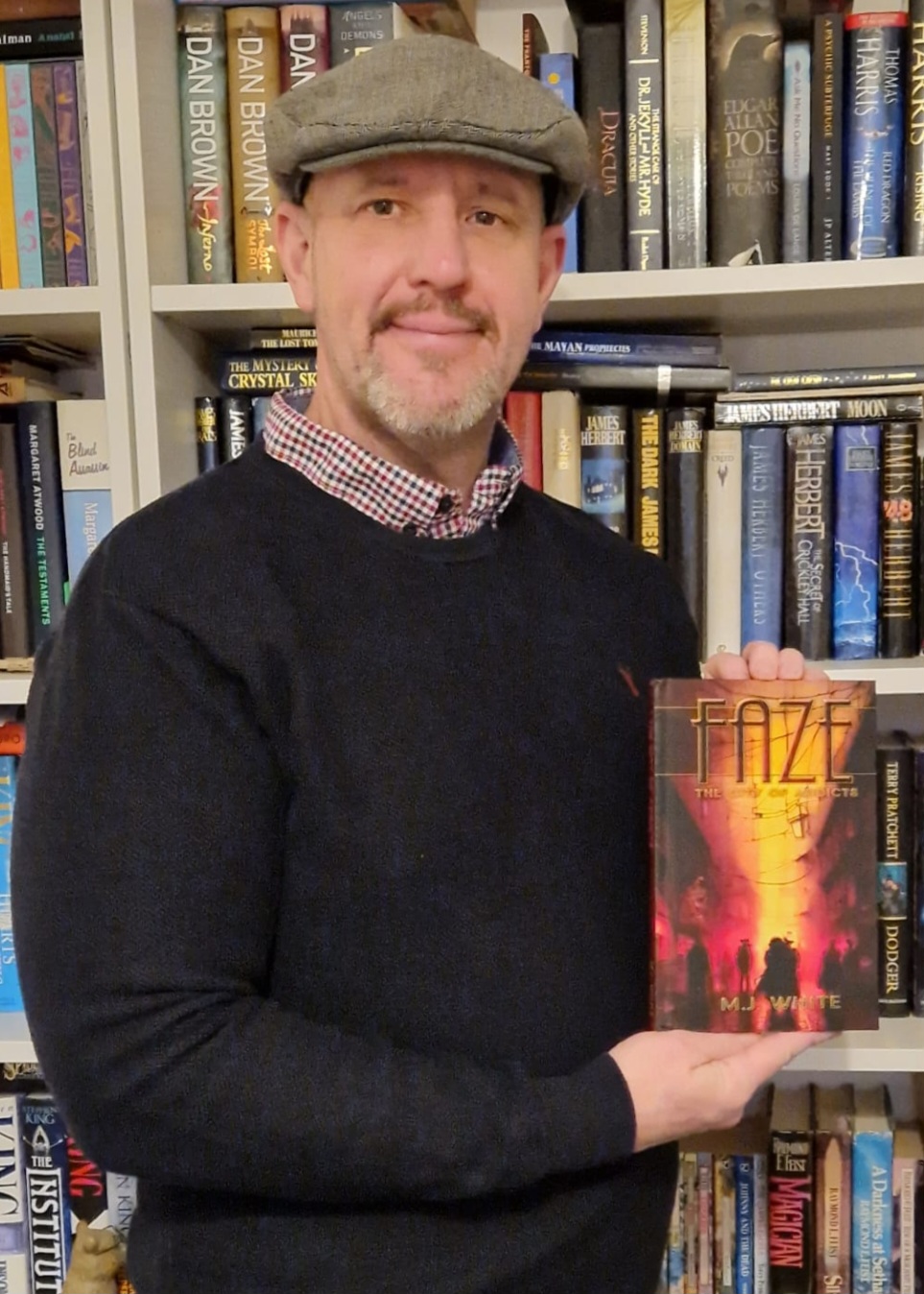 Man wearing flat cap holding up a book while standing front of a bookcase