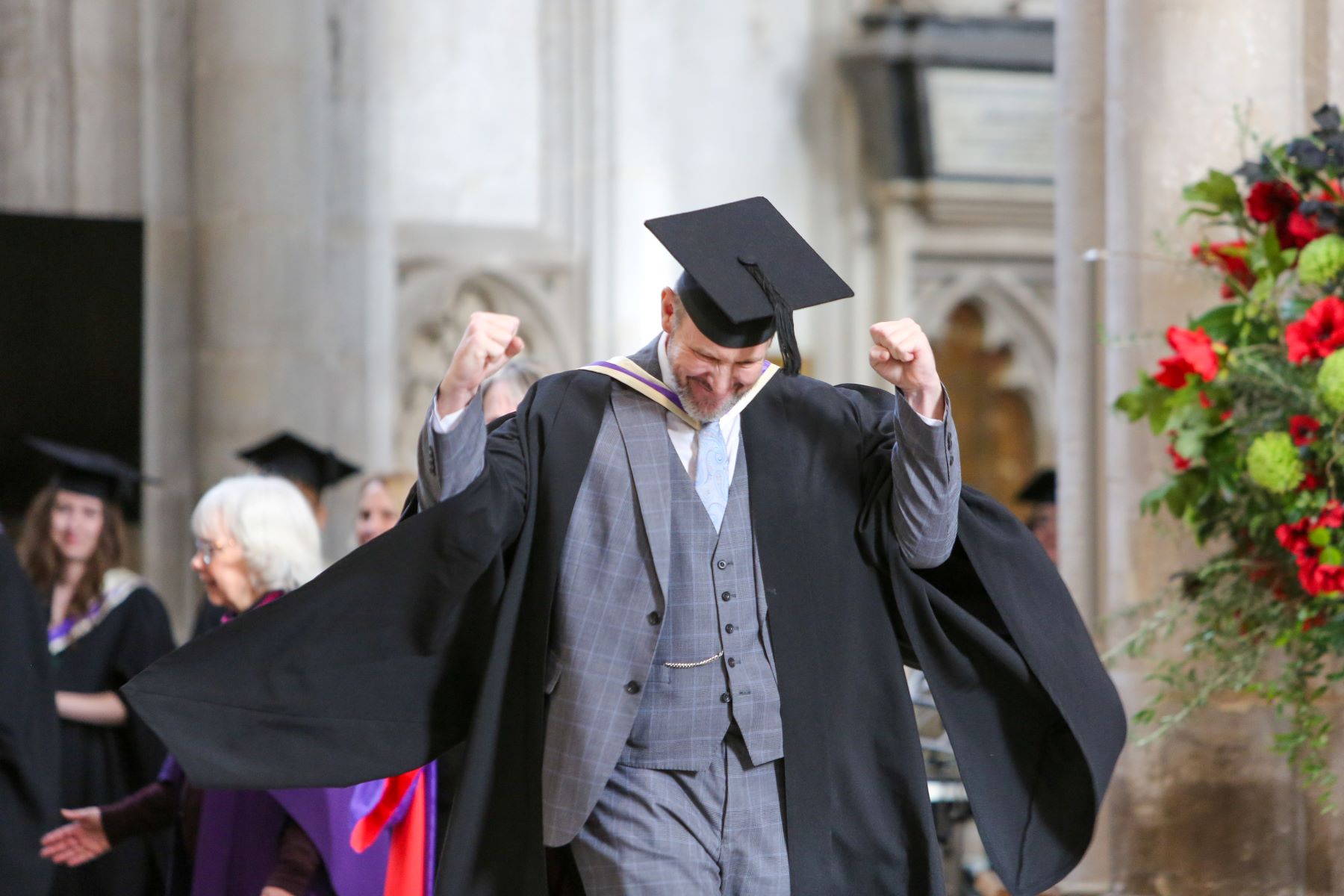 A mature student in suit, cap and gown celebrating with arms aloft