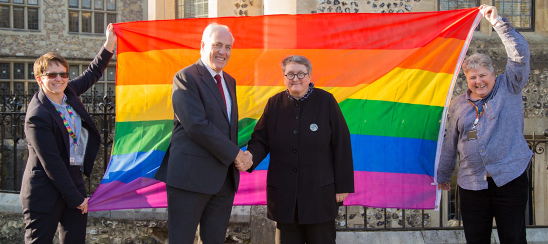 Two people shaking hands in front of a rainbow Pride flag