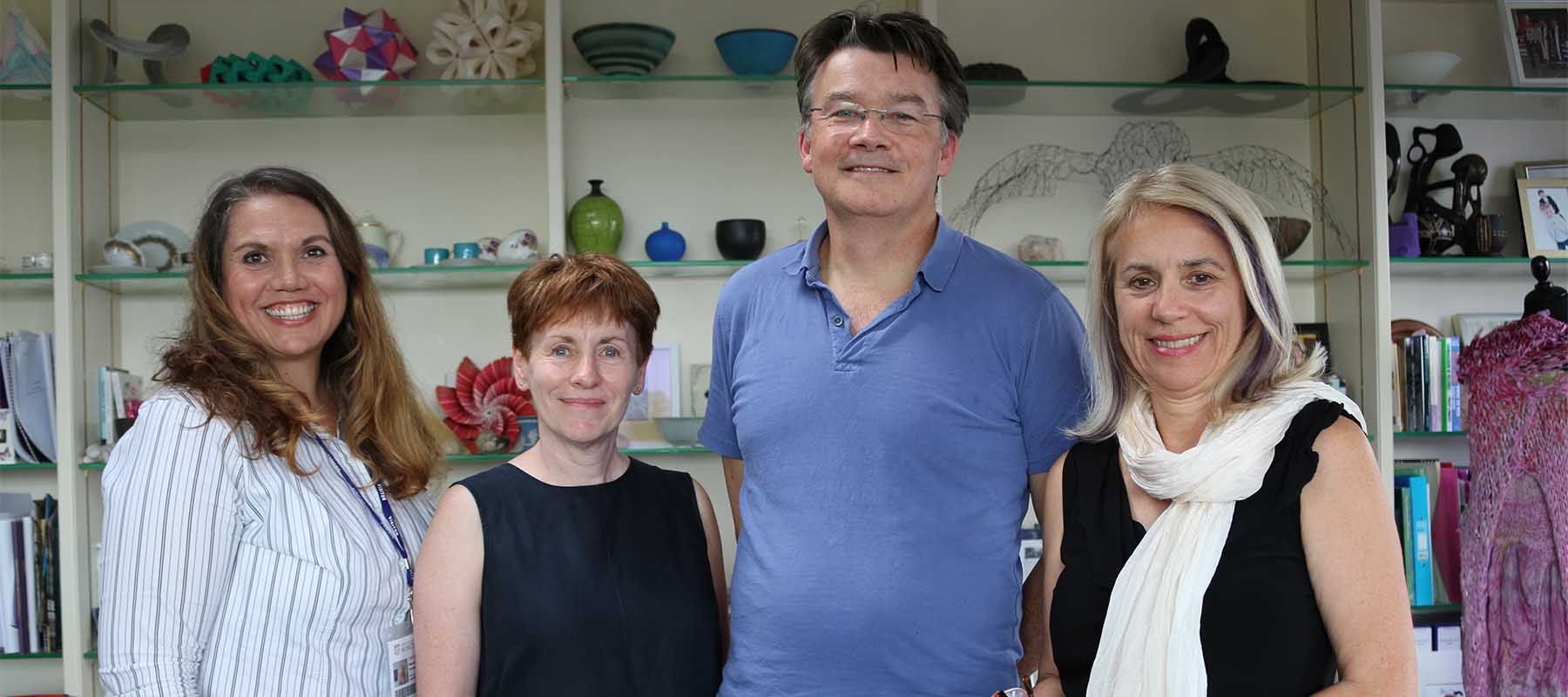 Man in blue polo shirt standing with three women in an office on front of bookshelves