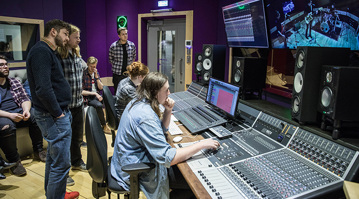 Man at the mixing desk in the recording studio watched by members of Alt-J 