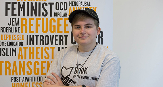 Woman representing 'Book' in human library stands in front of wall covered in labels like 'Feminist, OCD, Jew, Refugee, Introvert etc'