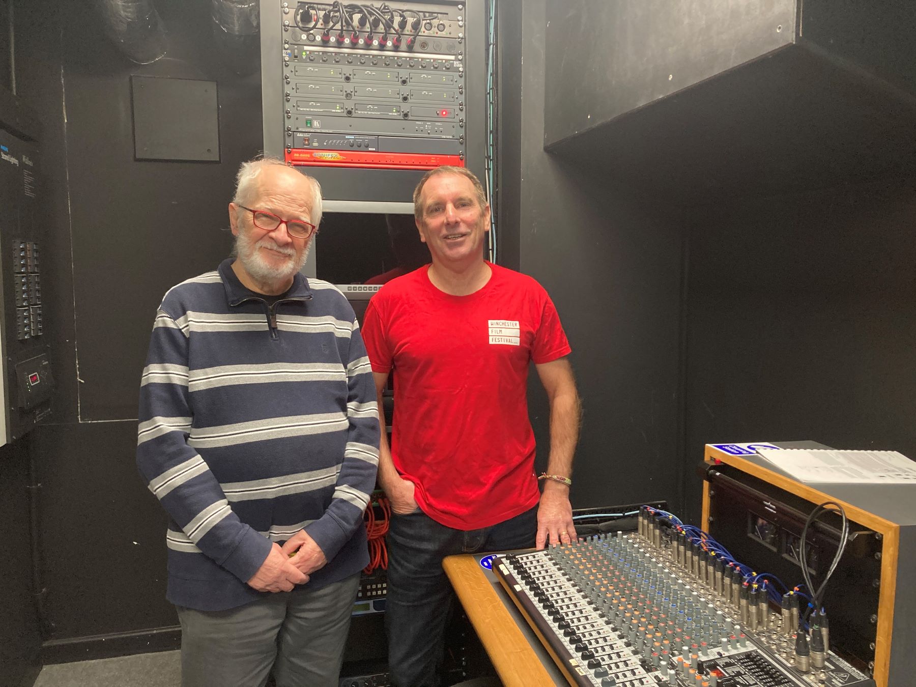 One older man with beard and glasses and a middle aged man in red t shirt next to mixing desk media studio