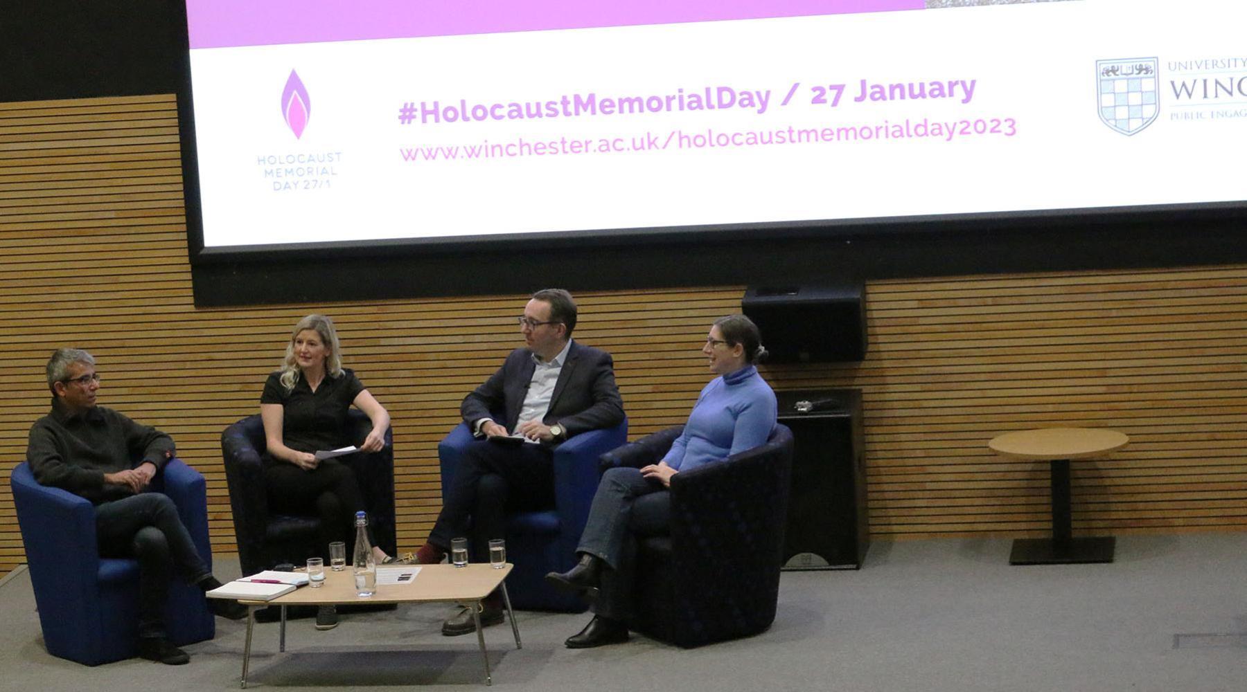 four people seated on a stage talking, in front of an image saying Holocaust Memorial Day 2023