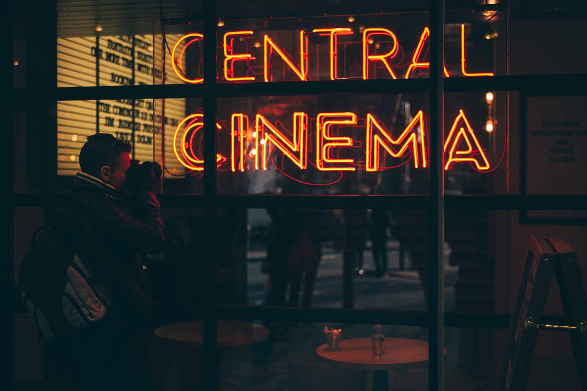 A person taking a picture of a cafe window which has a neon sign saying 'CENTRAL CINEMA'