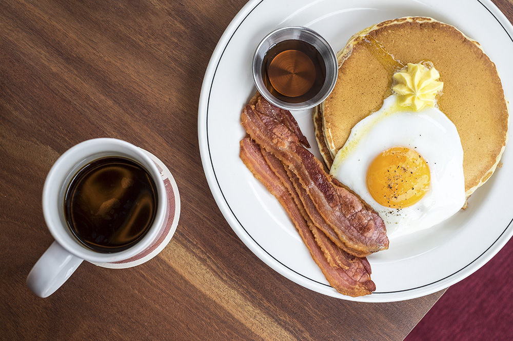 A breakfast plate containing pancakes, egg and bacon, with a mug of coffee